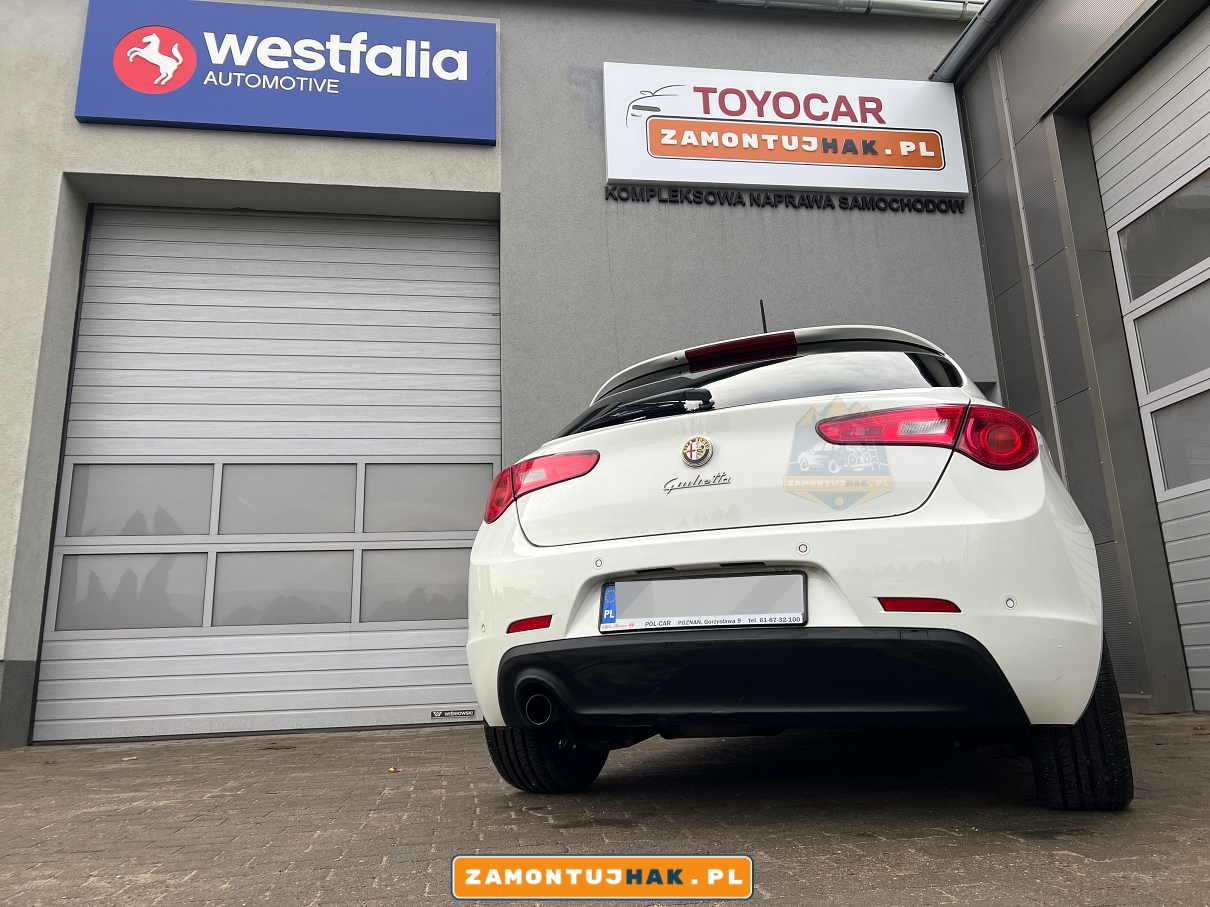 Alfa Romeo z hakiem holowniczym na tle warsztatu montażowego w Poznaniu: Obraz pokazuje Alfa Romeo z zamontowanym hakiem holowniczym, a w tle widać warsztat montażowy w Poznaniu, gdzie usługa została wykonana.